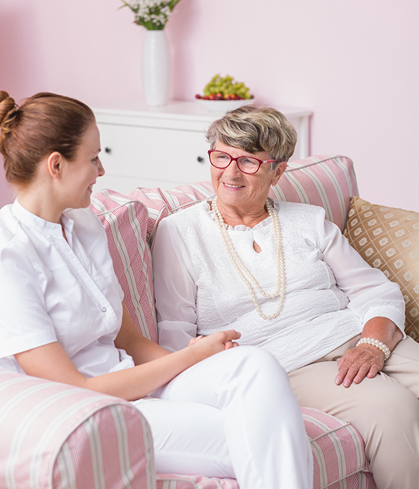 Caregiver and elderly woman smiling and talking while seated together on a couch in nursing home
